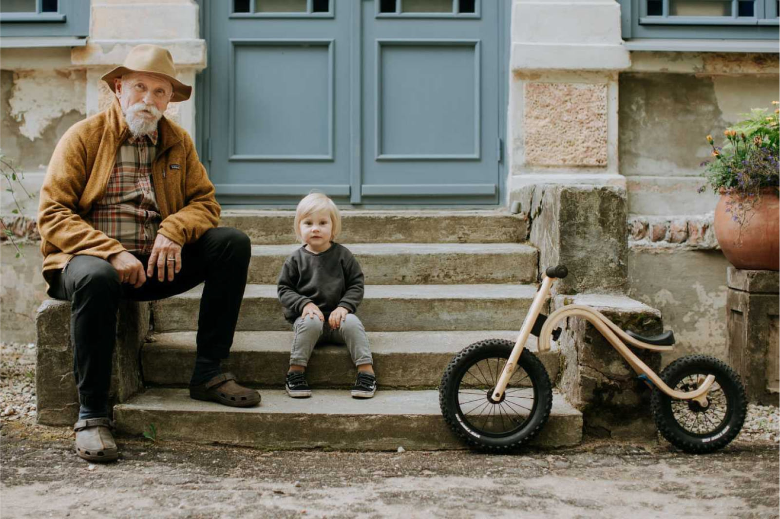 Leg&go brand wooden balance bike from Sostter marketplace beside a young child and older man sitting on stone steps outside a house
