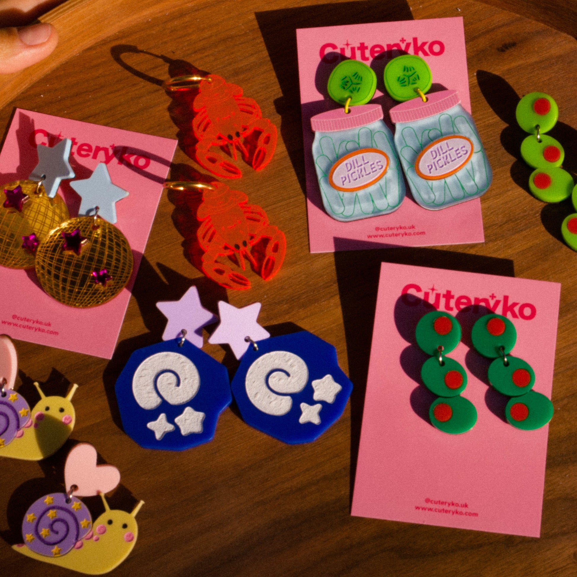 Colorful earrings on pink cards with 'Cutenyko' branding on a wooden surface.
