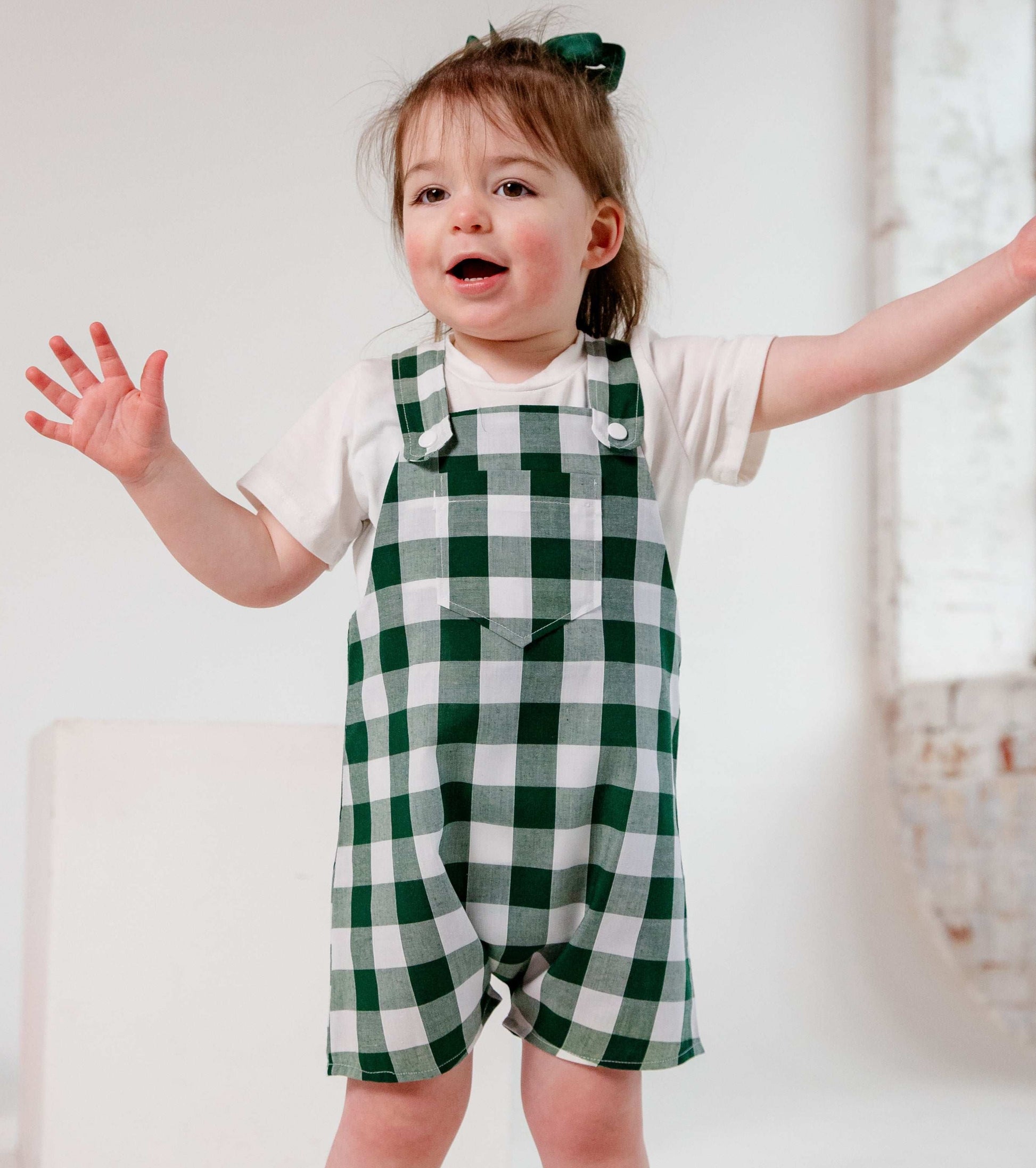 Child wearing green checkered overalls standing in a bright room with white walls and a brick wall.