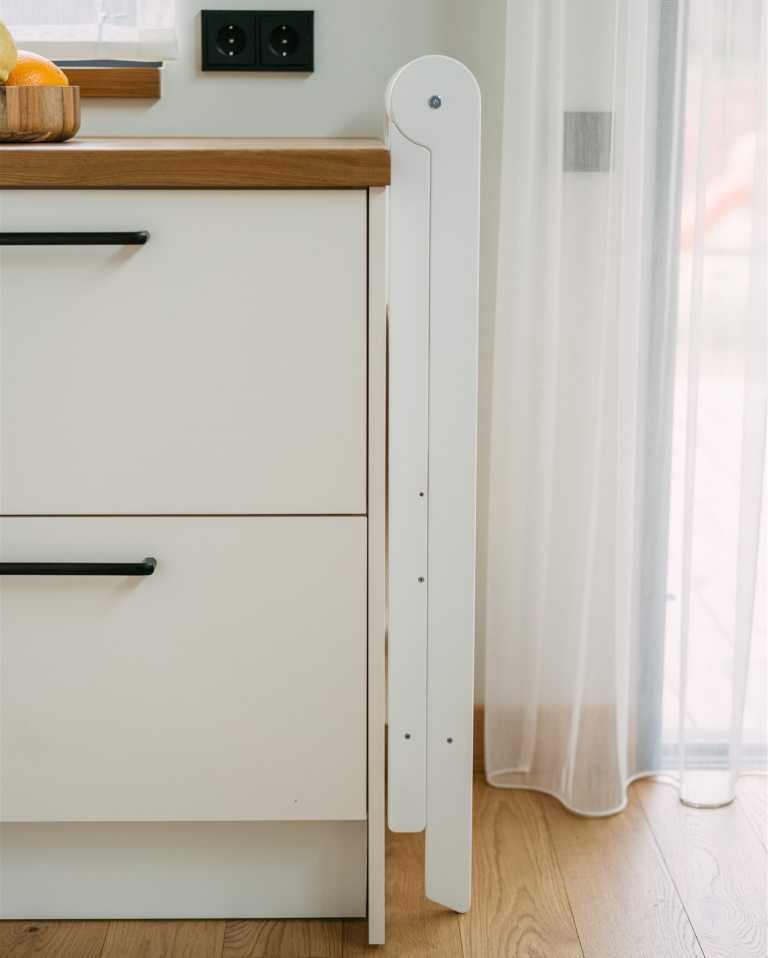 a child in a soft cotton outfit stands safely inside the foldable birch helper tower, reaching for a bowl on the counter
