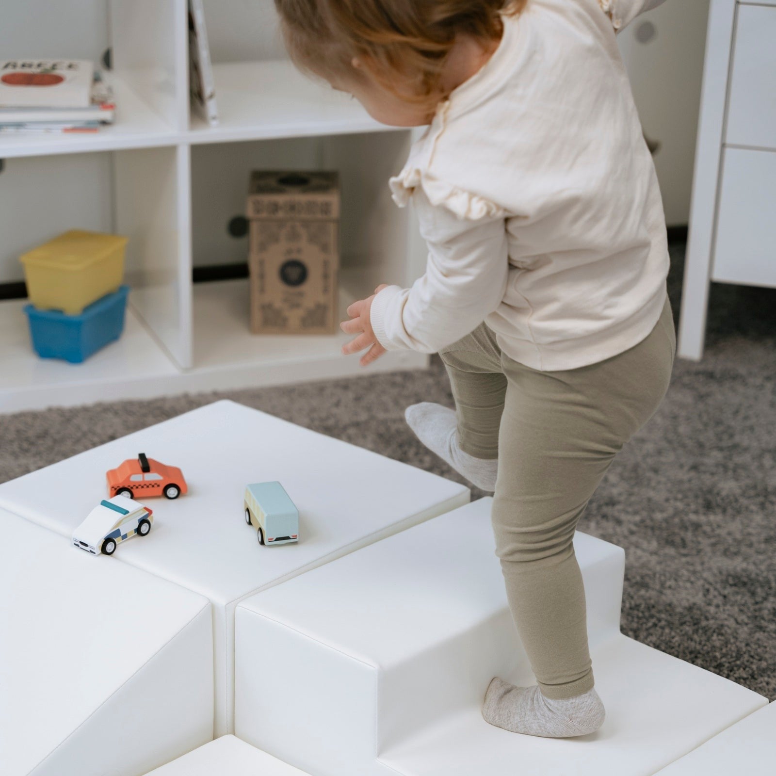 a child's hand reaching for a white foam corner climber block in a sunlit playroom