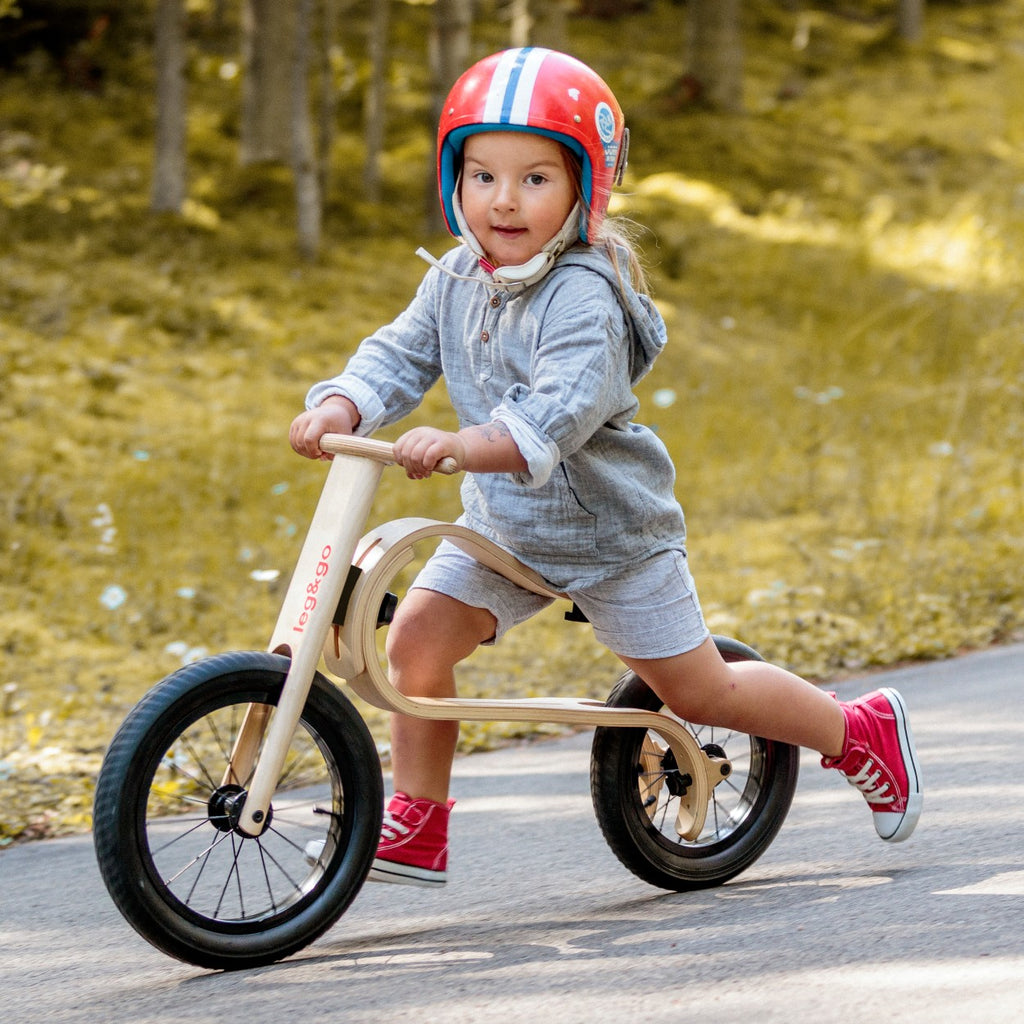 a bold toddler confidently astride a three-in-one wooden balance bike on a sunlit path