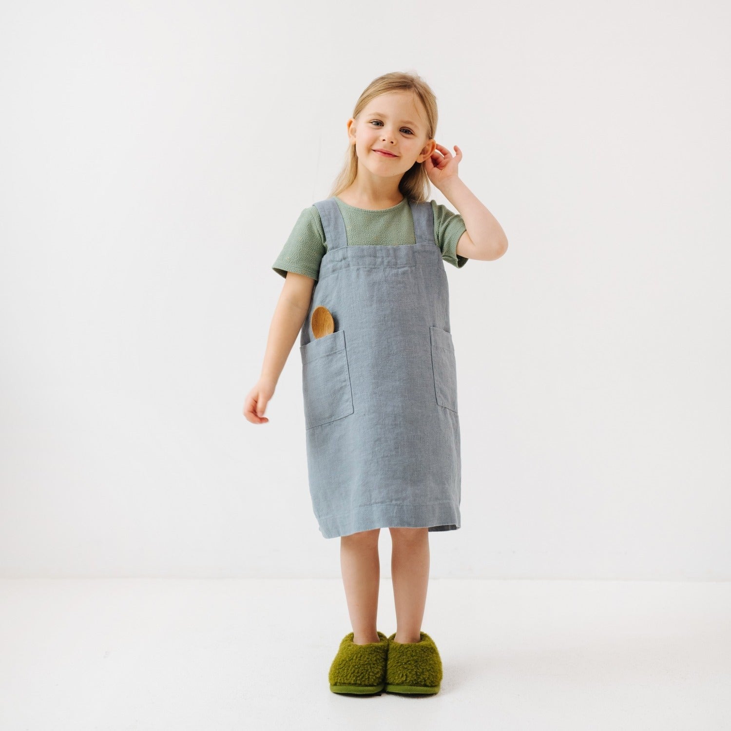 side view of a child in a soft blue fog linen apron, smiling proudly while holding a wooden spoon