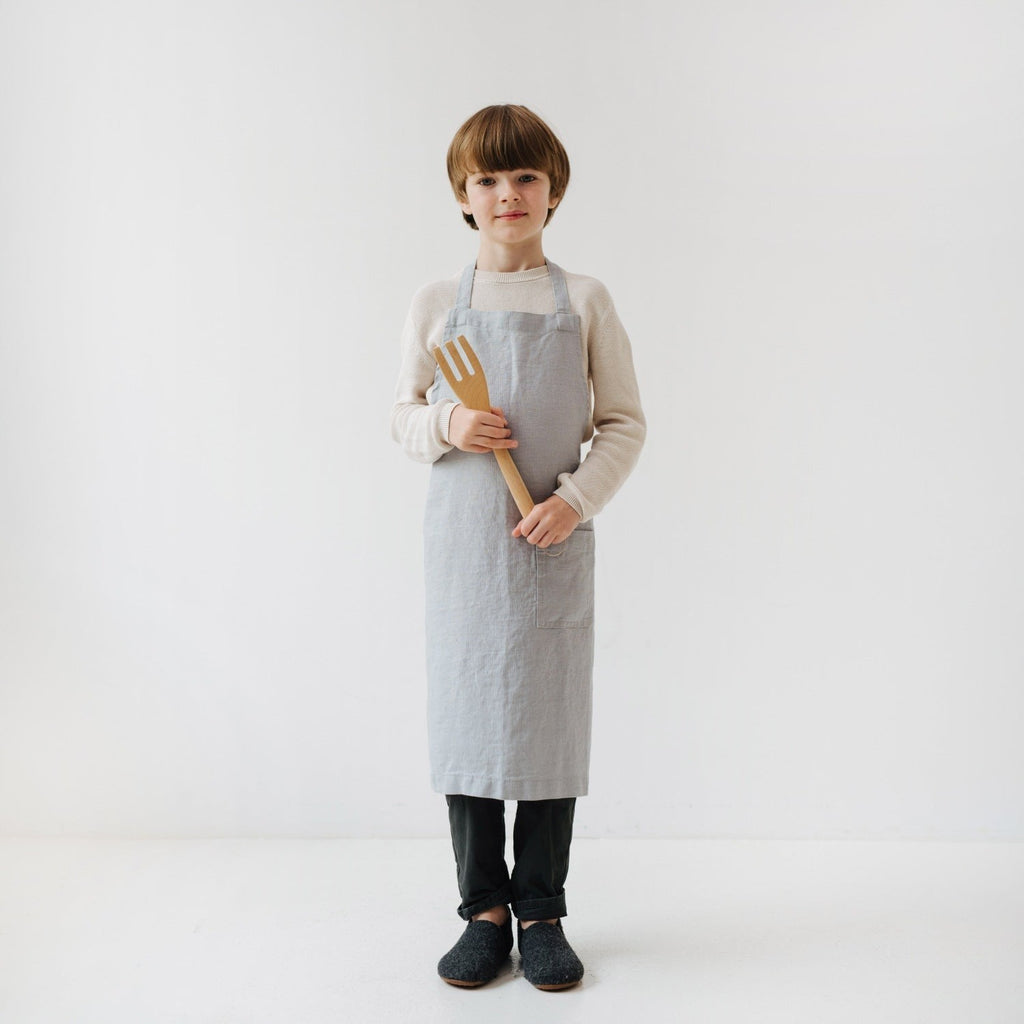 a playful kid in a light grey linen apron laughing while holding a wooden spoon
