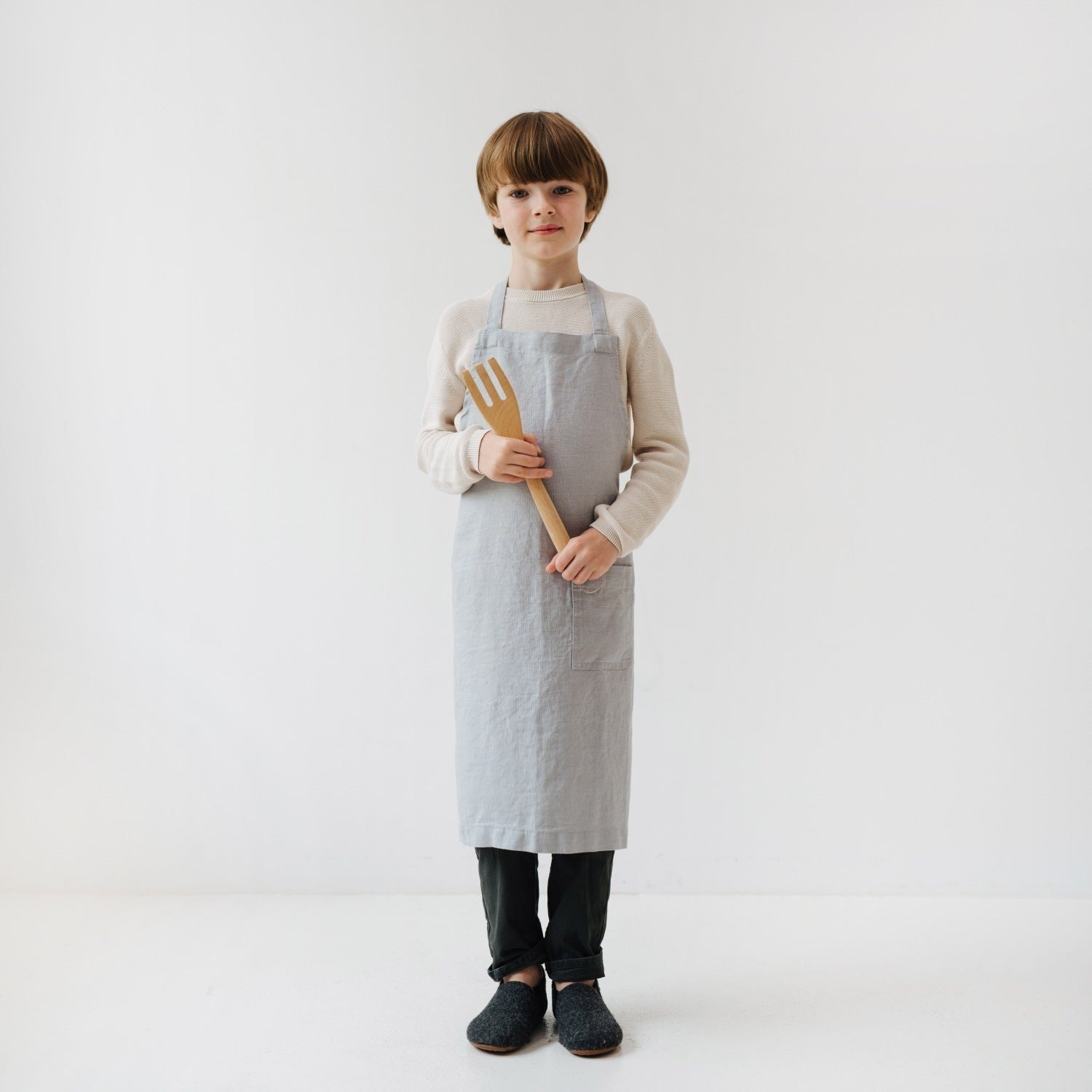 a playful kid in a light grey linen apron laughing while holding a wooden spoon