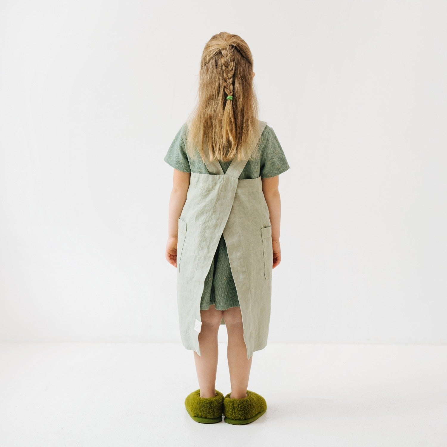 confident child in a sage linen pinafore apron standing in a sunlit kitchen