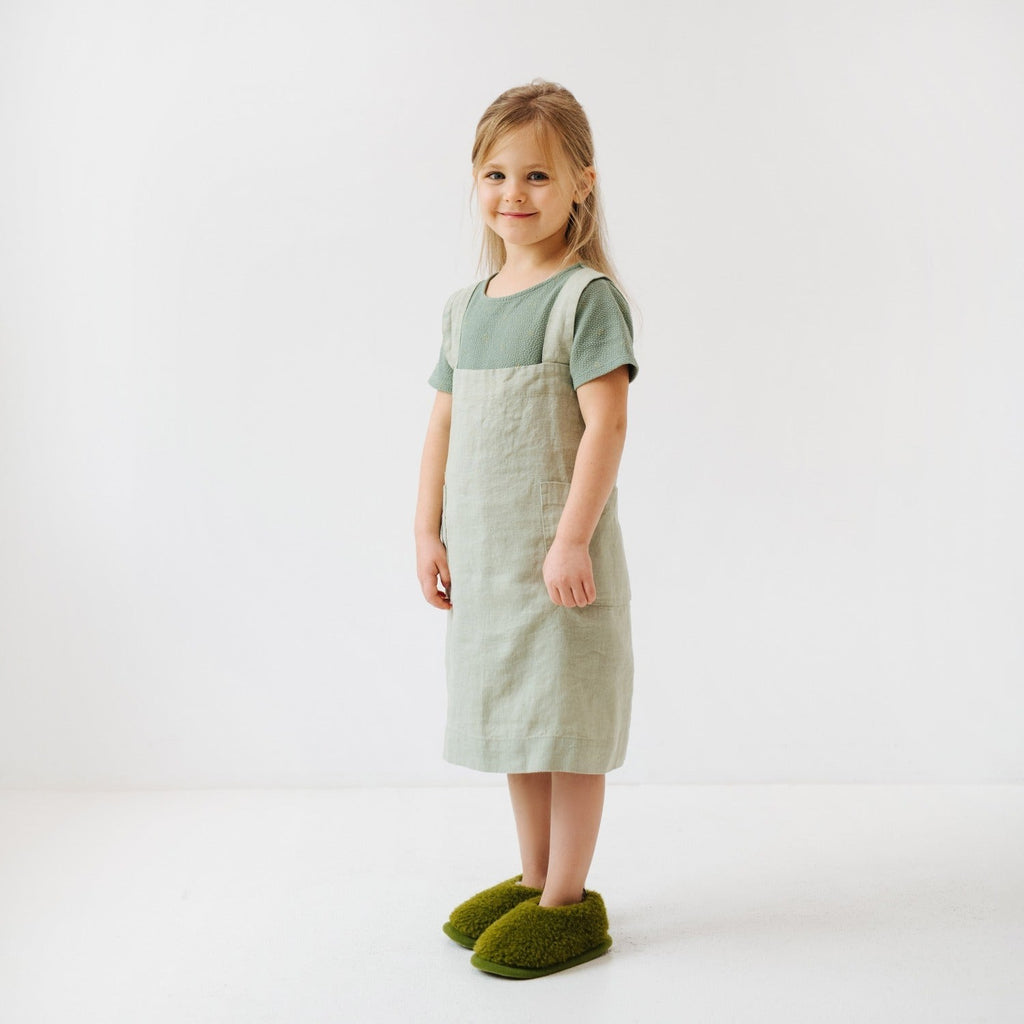 a child wearing a soft sage linen pinafore apron, smiling warmly in soft morning light
