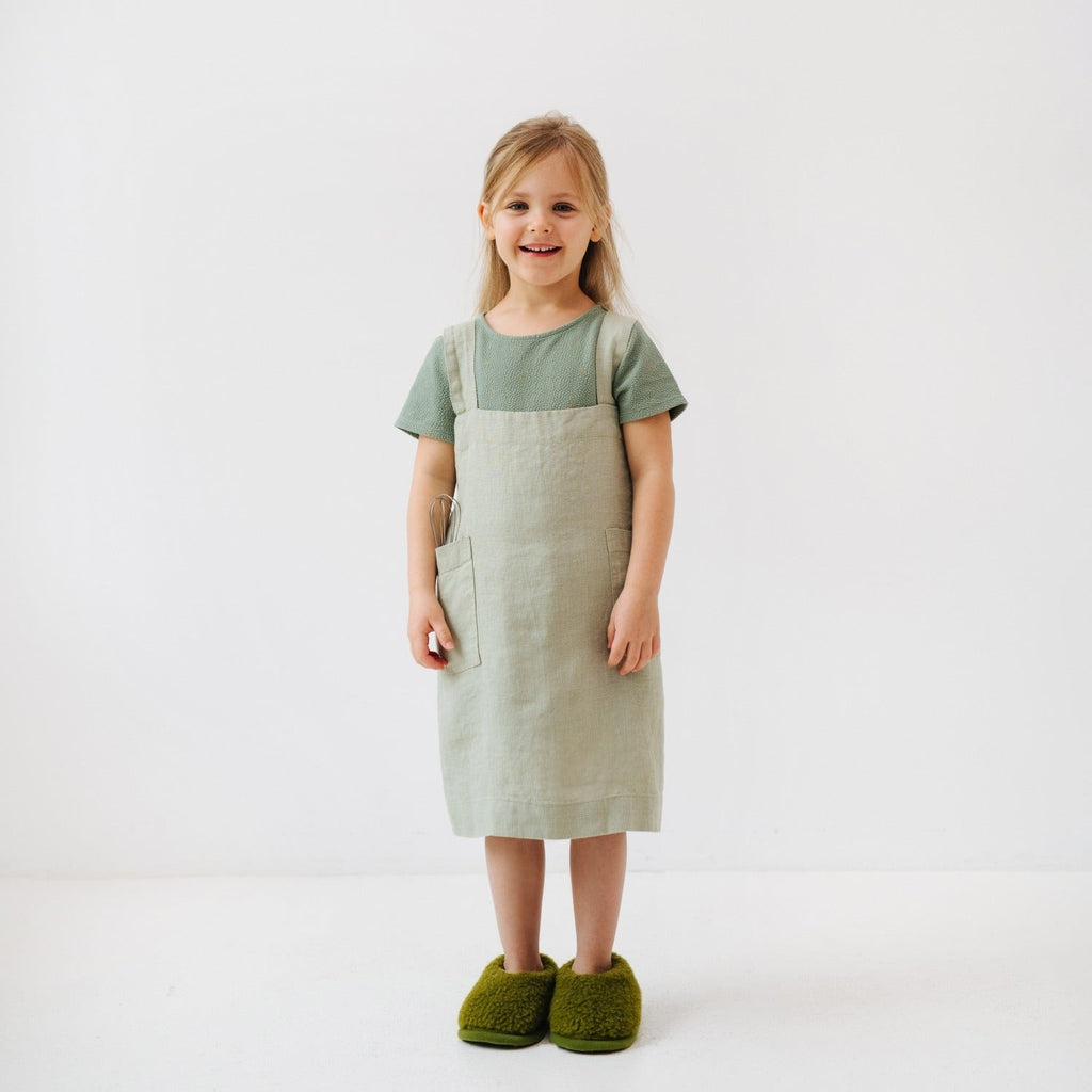 a child laughing while wearing a sage linen pinafore apron, hands in the pockets ready for a baking adventure