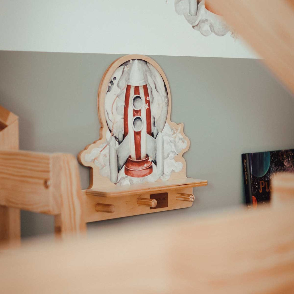 a playful rocketship wall shelf with a dinosaur sticker and three pegs for hanging small items