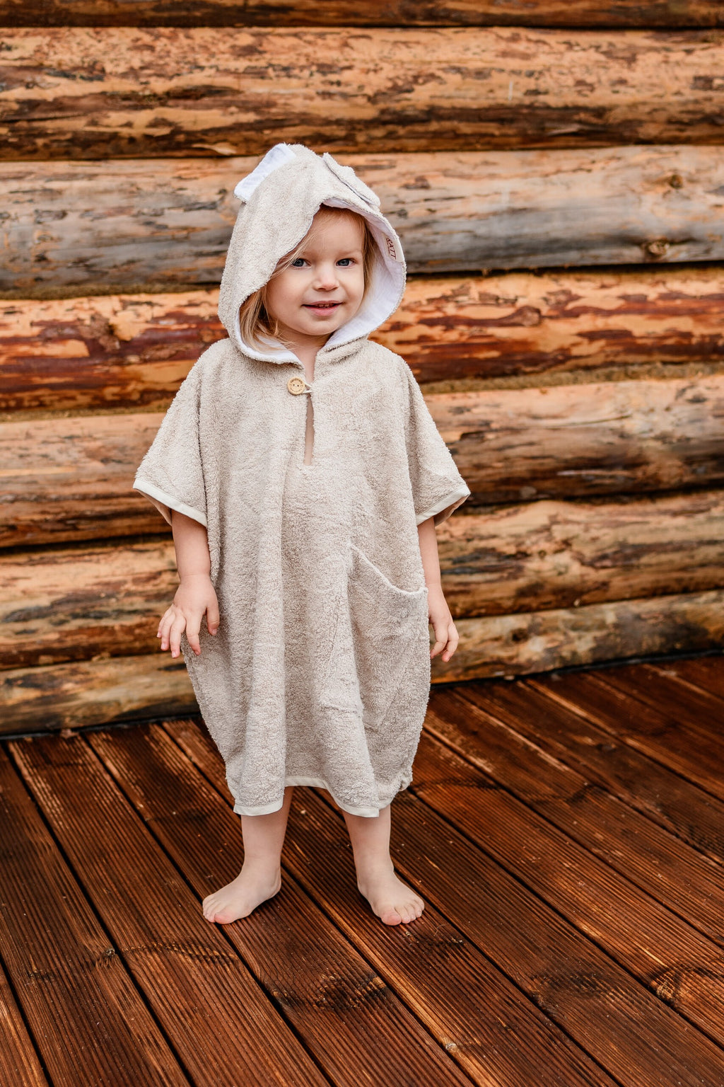 a toddler wrapped in a soft bamboo poncho, smiling warmly with the hood up in gentle light