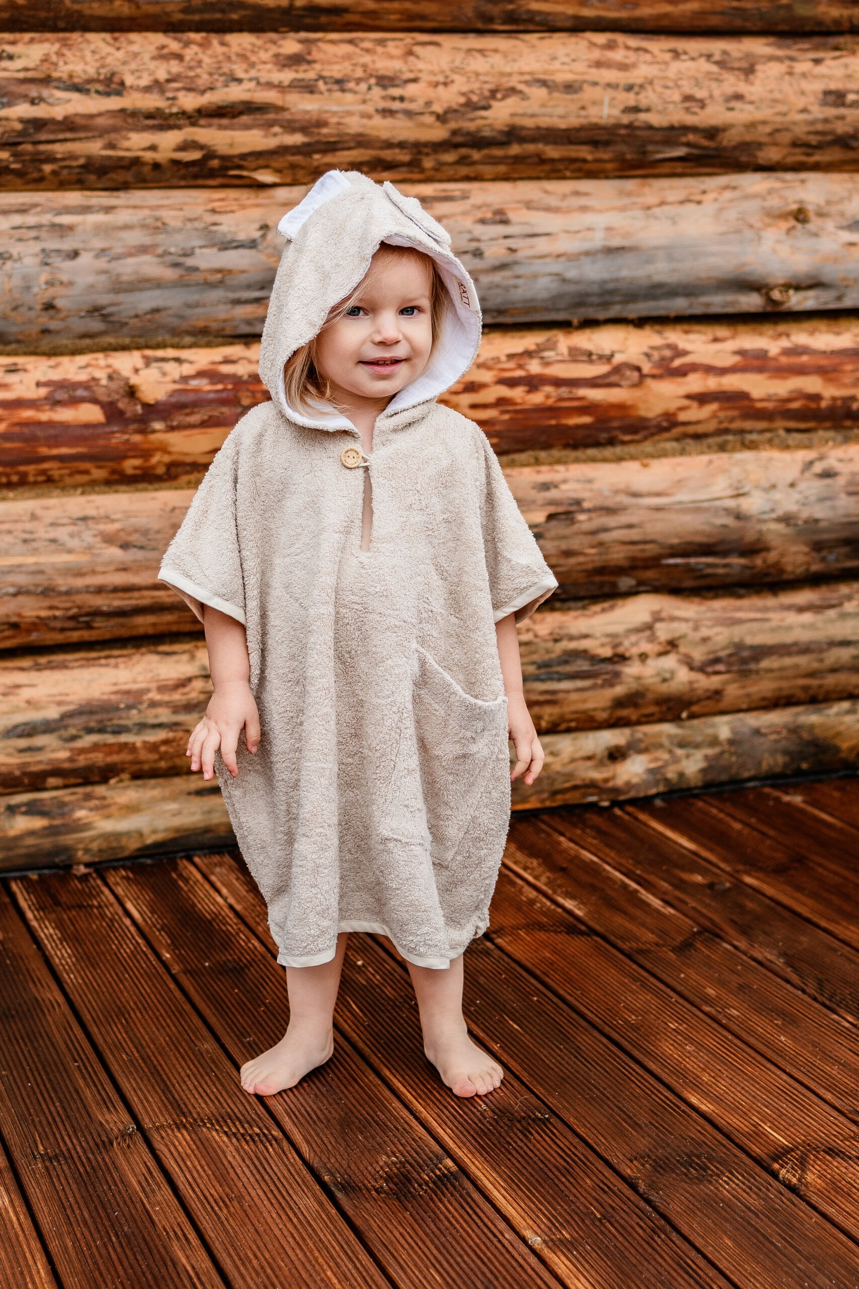 a toddler wrapped in a soft bamboo poncho, smiling warmly with the hood up in gentle light