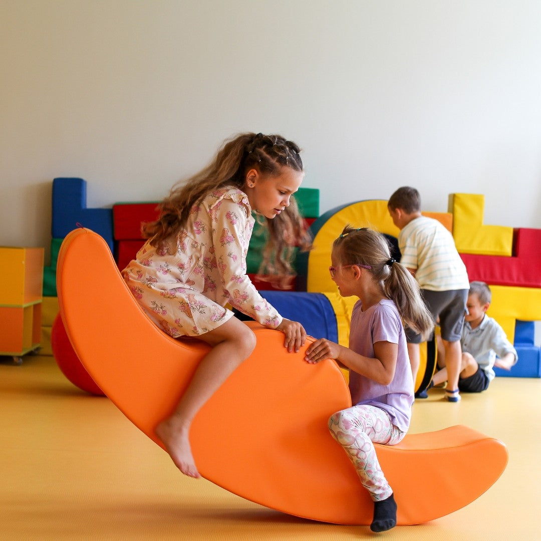 two toddlers laughing on a beige double rocker in a sunlit playroom