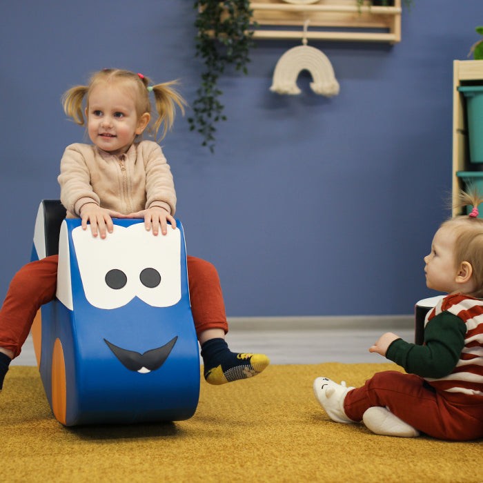 smiling toddler gleefully riding a soft blue car toy across a sunlit playroom floor