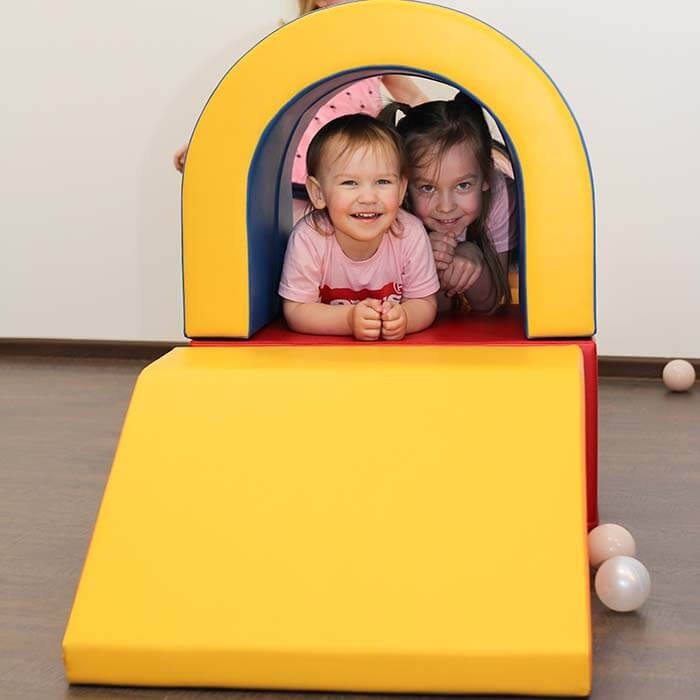 a child peeking out from a vibrant multicolor soft play tunnel with a big smile