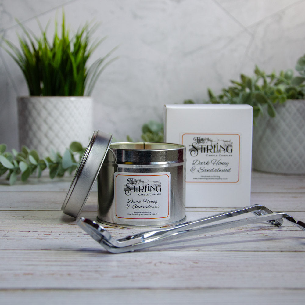 Silver candle with a label and packaging on a wooden surface with plants in the background
